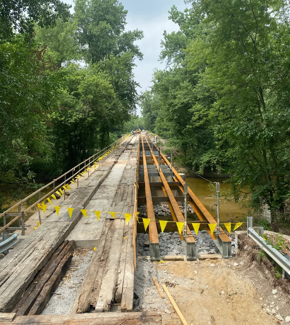 A partially constructed bridge made of wood and metal spans across a small river, surrounded by trees. Yellow caution flags hang along the sides.