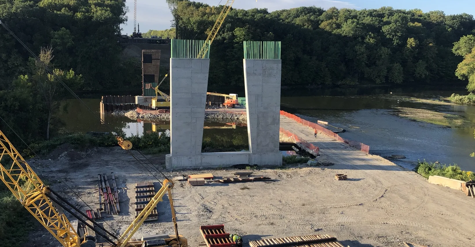 Construction site with two large concrete pillars by a river. Cranes and materials are visible around, with trees in the background.