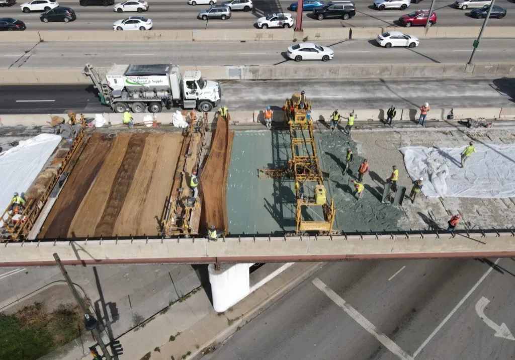 Aerial view of a bridge under construction with workers and machinery. Traffic is visible on adjacent roads.