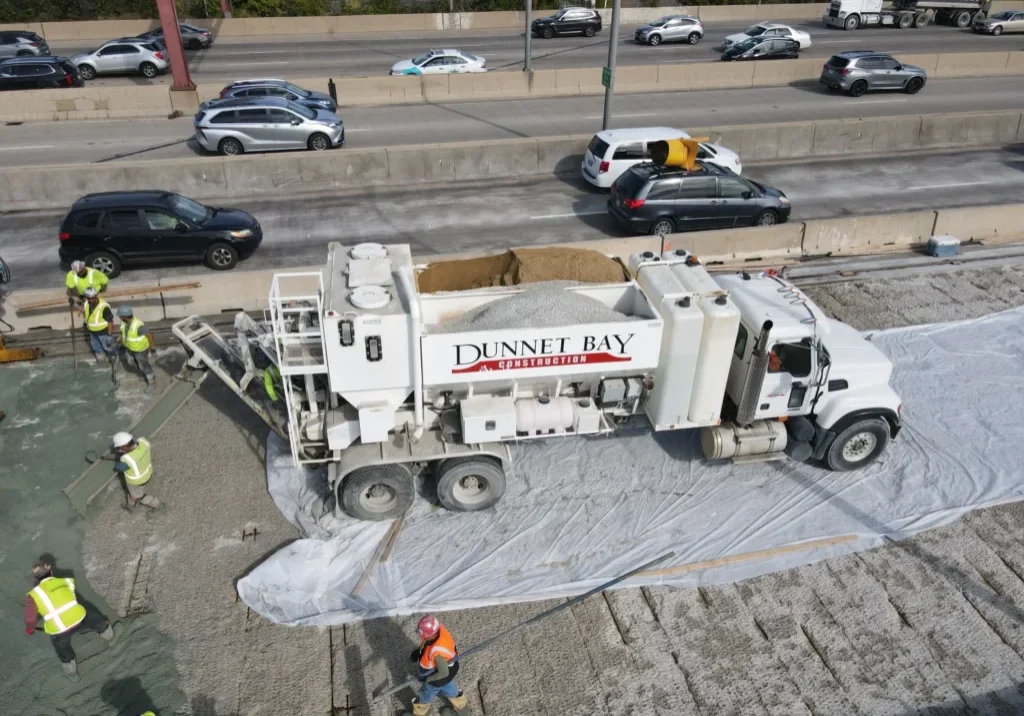 Construction workers operate near a large cement truck on a highway. Traffic moves slowly on the opposite side. Protective tarps cover sections of the road.