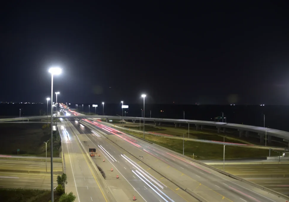 A long-exposure night shot of a highway with light trails from vehicles, against a dark sky, and illuminated by overhead streetlights.